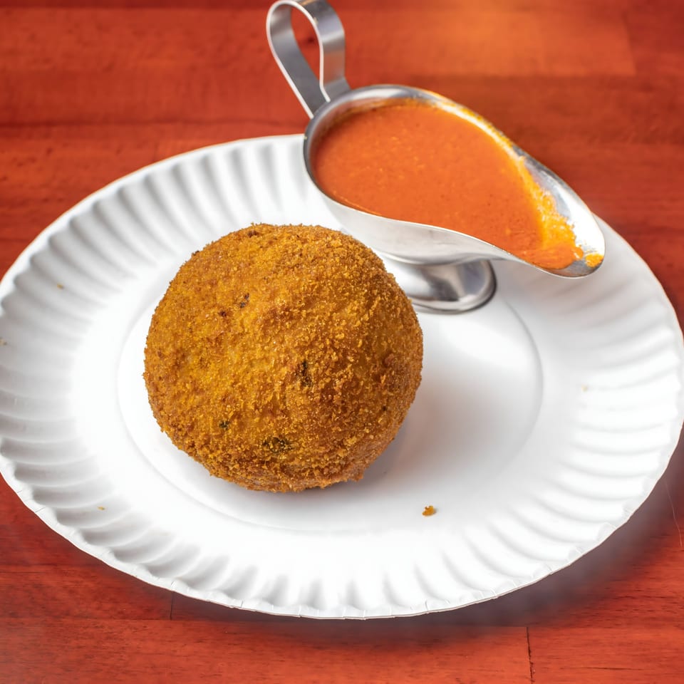 A golden-brown arancini ball on a white plate with a silver gravy boat of red sauce.