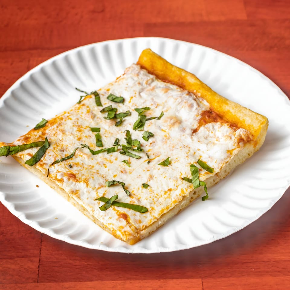 Square slice of Villa Formia's white pizza with ricotta, mozzarella, and fresh basil on a paper plate.
