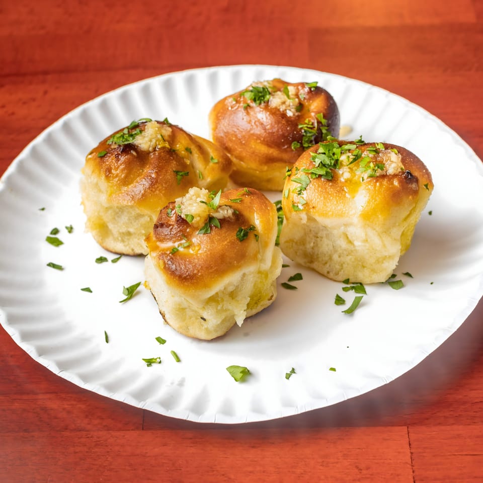 Four golden-brown garlic knots, topped with minced garlic and fresh parsley, on a white plate.