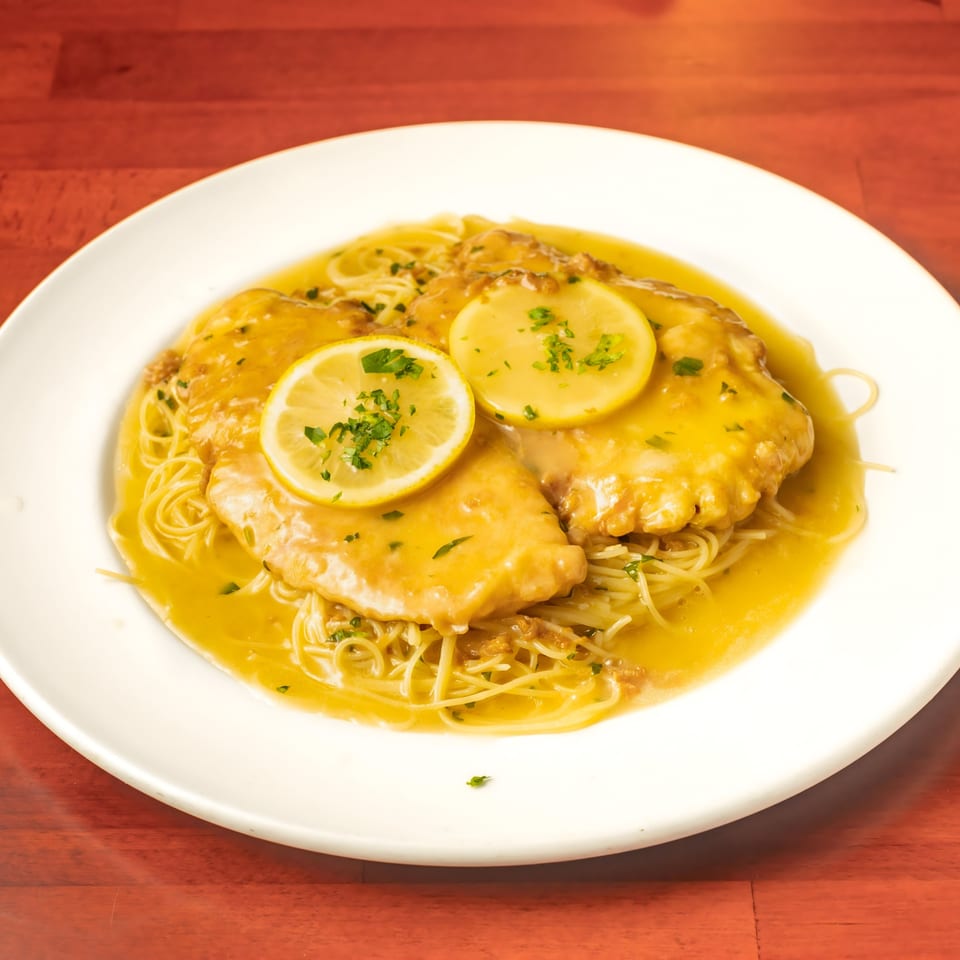 Chicken piccata with lemon slices and parsley over spaghetti, in a rich sauce on a white plate.