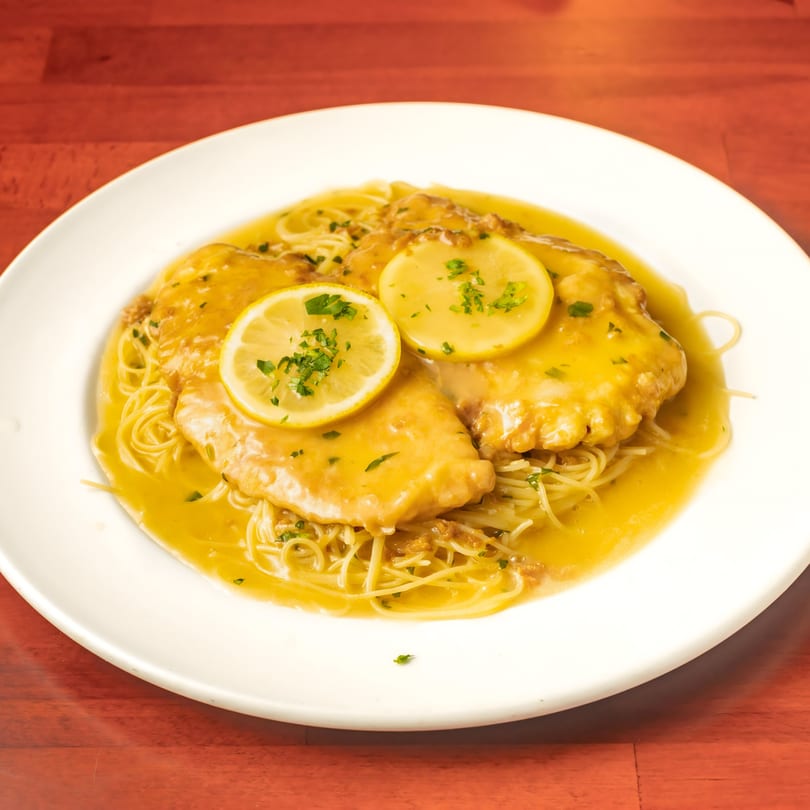 Chicken piccata with lemon slices and parsley over spaghetti, in a rich sauce on a white plate.