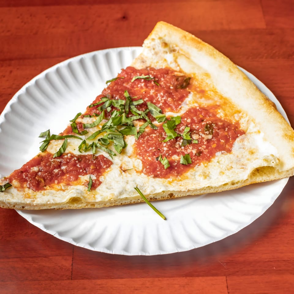 A slice of Villa Formia pizza with red sauce, white cheese, and fresh basil on a paper plate.